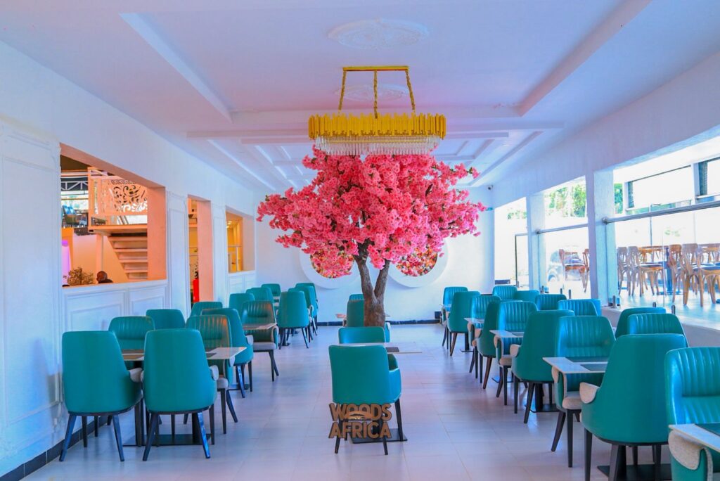 A bright dining room at Woods Africa shows teal chairs, white walls, and a tall pink blossom tree beneath a gold chandelier.