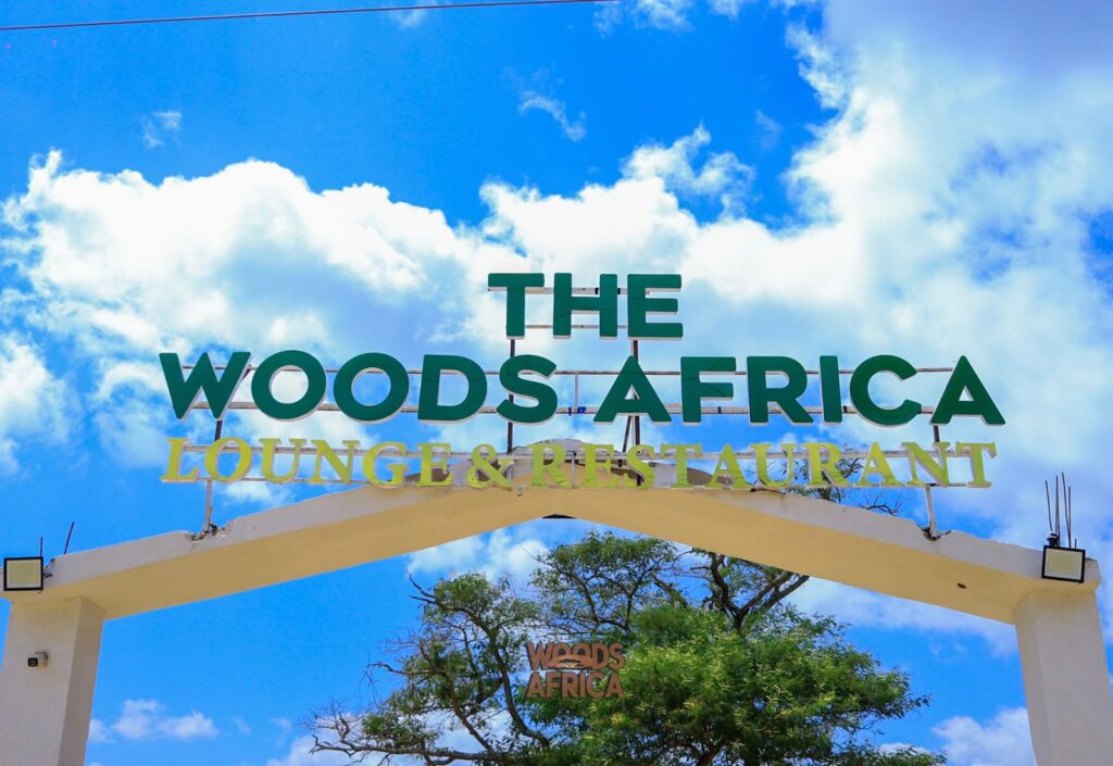 The Woods Africa Lounge & Restaurant entrance sign rises above a white arch against a vivid blue sky with scattered clouds.
